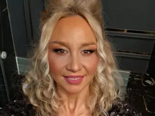 Smiling woman with blonde wavy hair and glamorous makeup wearing a black sequined outfit in a dark setting.
