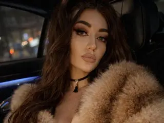 MiaBlackXO poses in a car wearing a black choker and a thick, tan faux fur coat, showing her long dark hair and heavy eye makeup.