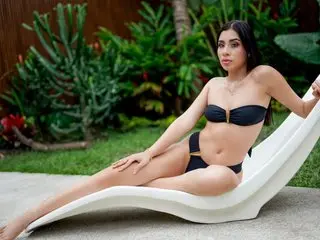 MiaColucci_GB, a woman with long dark hair, poses naked from the waist up while sitting on a white lounge chair outdoors.