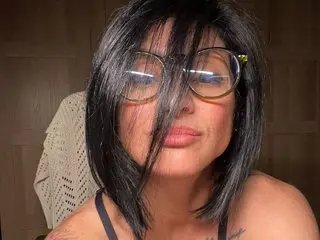 MiaFit poses with a direct gaze, wearing large glasses and showing her dark hair and bare shoulders against a plain brown wall.