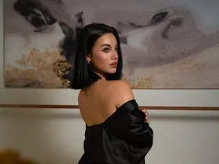 MiaGreenwood poses with her black hair down, showing her bare shoulders and back while wearing a black satin off-the-shoulder top against a neutral background.