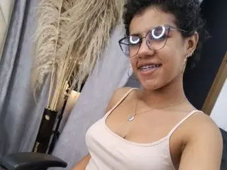 MiaPaytonnn with curly hair wearing a white tank top, smiling while posing in a room with pampas grass decor.