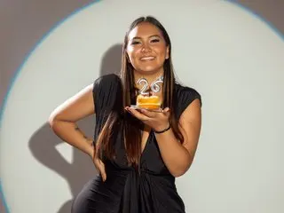 Mia_Laurent smiles while wearing a black sleeveless top, holding a small dessert or treat in her hand against a light grey background.