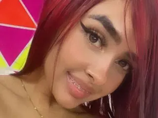 Mia_doll_66 smiles close-up, showing her face, dark eyes, and red hair against a bright, colorful background.