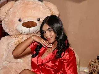 Smiling young woman in red satin pajamas posing next to a giant teddy bear in a cozy indoor setting.
