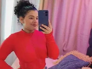 Miaa_Rous poses with her dark curly hair up, wearing a red long-sleeved top against a soft pink curtain background.