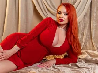 MickyBells with long red hair poses in a tight red dress while reclining on a gold satin sheet.