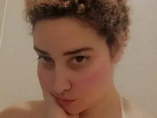 MielMal looks at the camera with a soft expression, showing her face and curly hair against a plain wall.