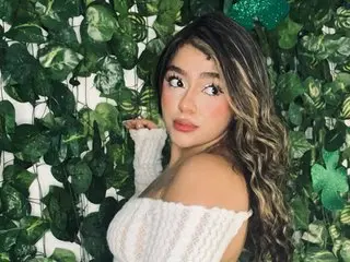 Miia_Sinner poses with her long wavy hair down, wearing a white off-the-shoulder top against a lush green leaf background.
