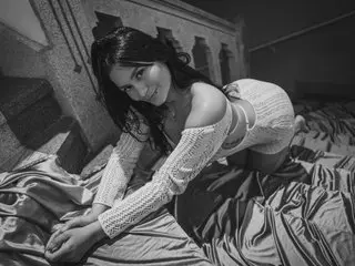 Miiaa_jones poses naked in a white lace top, leaning forward over rumpled bed sheets with her dark hair falling over her shoulders.