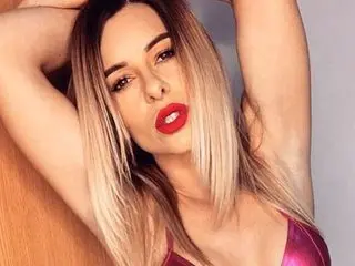 MikaelaWittBabestation poses with her hands behind her head, wearing red lipstick and a shiny pink bra.