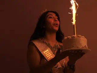 Milan_Rousse poses with a lit birthday candle, wearing a silver tiara and a decorative sash against a dark background.