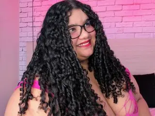 MileyCollinss smiles with long, dark curly hair while wearing a pink top in a room with pink brick walls.