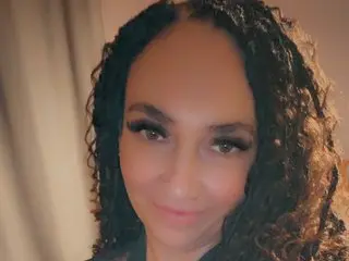 MilfMillie poses with a close-up view of her face and long, curly brown hair against a plain background.