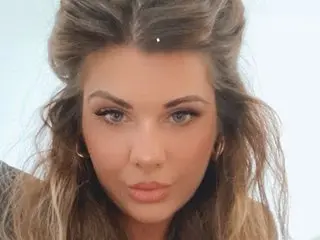 Milf_Sutton poses with her long brown hair framing her face, looking directly at the camera with a neutral expression against a plain white background.