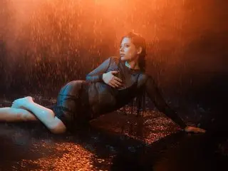 Moody portrait of a woman in a black bodysuit reclining in warm orange rain under dramatic lighting.