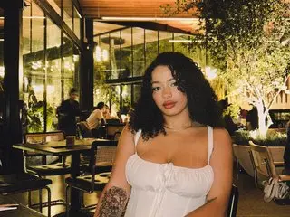 MinaKyoto poses with her dark wavy hair, wearing a white tank top while sitting at a table in an outdoor restaurant setting.