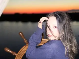 Minatyan18 poses with her long hair falling over her shoulder against a sunset background over a body of water.