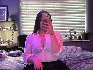 MiriXxX sits on a bed in a white button-down shirt, taking a mirror selfie in her bedroom.