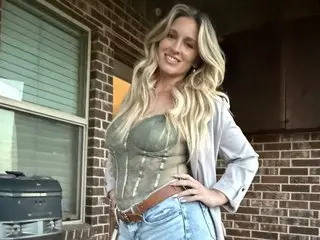 MissBehavin90 poses outdoors in a green corset and light blue denim, showing off her long blonde hair and hand on her hip.