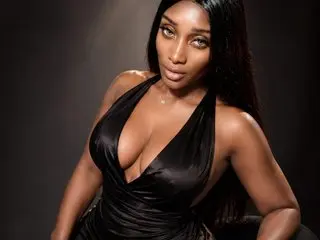 MissColette poses in a low-cut black satin dress, showing deep cleavage and her long dark hair against a dark background.