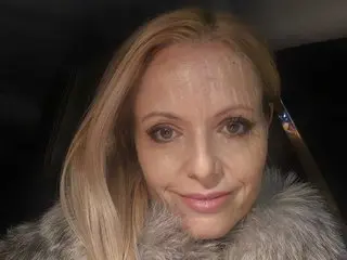 MissJazmin poses with her blonde hair down, wearing a thick, grey faux-fur collar or coat against a dark background.