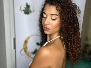 MissMilano_xo poses with her long, curly brown hair down, showing her bare shoulders and chest against a white background.