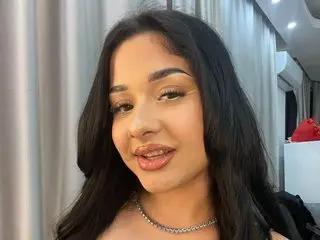 MissNaomiBelle poses with long dark hair and a silver chain necklace, looking directly at the camera with a soft smile.