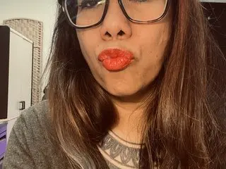 MissZeena pouts her lips with red lipstick, wearing glasses and a grey top, with her long brown hair framing her face.