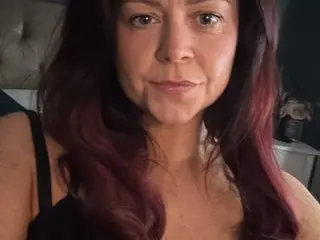 Portrait of a woman with long reddish-brown hair wearing a black strap top in a dimly lit bedroom setting.