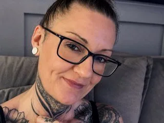 Missyinkxxo smiles while wearing black-rimmed glasses and a black tank top, showing her dark hair pulled back and detailed neck tattoos.
