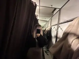 MistressLouUK poses with her blonde hair, holding a black smartphone up to her face in a room with light-colored walls and hanging brown fabric.