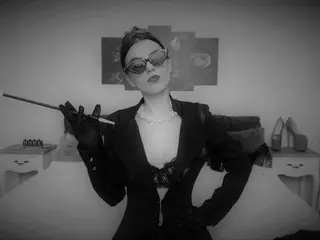 MistressMadelyn poses in a black jacket and dark sunglasses, wearing a silver necklace and holding a thin wand against her chest.