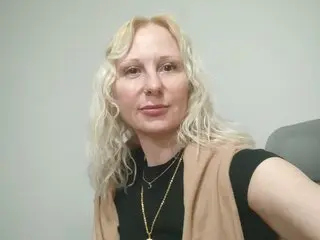 MistressParisParker poses with her long blonde hair down, wearing a black top and tan vest against a plain grey wall.