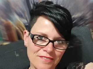 MollySun wearing black glasses and looking directly at the camera with short black hair, posing in a dark setting.