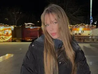 MollyyMay poses with long blonde hair wearing a black puffer jacket against a dark outdoor background with warm lights.
