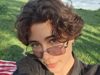 MorganMiiler poses with a soft smile, wearing tinted sunglasses and a black shirt, with her wavy brown hair framing her face against a grassy outdoor background.