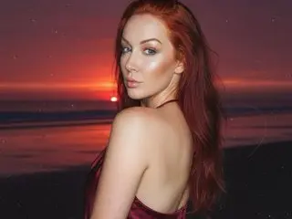 MorganReigns poses with her long red hair, showing her bare shoulder and back against a sunset beach background.