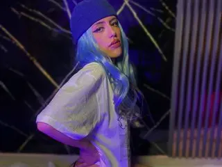 MorganaSmitt poses in a white t-shirt and blue beanie, showing her blue hair and face against a dark, purple-lit background.