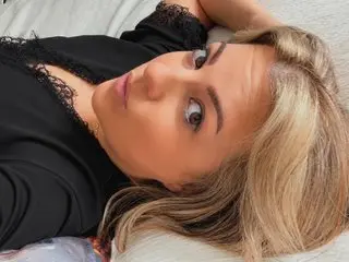 MsBelleWarick poses close-up, showing her face and blonde hair while lying back against a white surface in a black lace top.