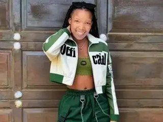 MsDareAssDevil smiles while wearing a white and green zip-up jacket and matching green shorts, showing her hair in long braids.