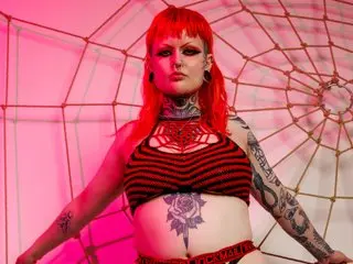 MxSalemSage poses with red hair and heavy tattoos, wearing a red striped crop top against a red spiderweb background.