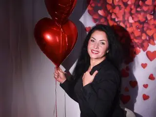 Smiling woman in a black outfit holding red heart balloons against a romantic Valentine's Day backdrop.