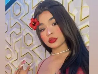 Young woman with a red hibiscus flower in her hair and red lipstick posing against a geometric patterned wall.