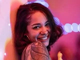 NaiadRae smiles while showing her face and dark hair, posing close up against a soft pink background.