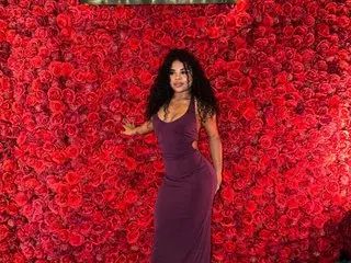 NaiaraReina poses with her long dark hair down, wearing a tight purple dress against a wall of red roses.