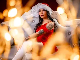 Naila_Mistress poses in a red lace bodysuit, showing her cleavage and legs while sitting on a white chair against a warm, blurred background.