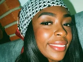 Smiling Black woman wearing a patterned headwrap and glowing makeup in a cozy indoor setting.