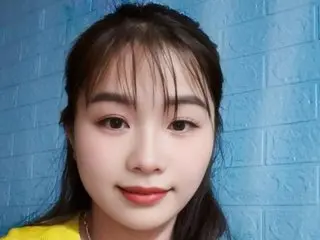 NaneyCen looks at the camera with long dark hair and red lipstick, wearing a yellow top against a blue brick wall.