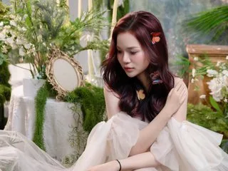 Naomi040 with long reddish hair sits amidst white flowers and greenery wearing a sheer cream-colored dress.